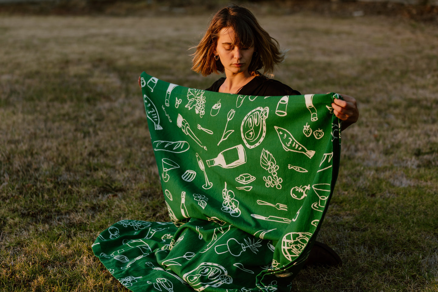 A woman holds the folded "Green Feast" tablecloth, showcasing its vibrant green color and minimalist white illustrations. The tablecloth, inspired by a 2023 trip to Greece, reflects a timeless elegance and simplicity, perfect for any dining setting.
