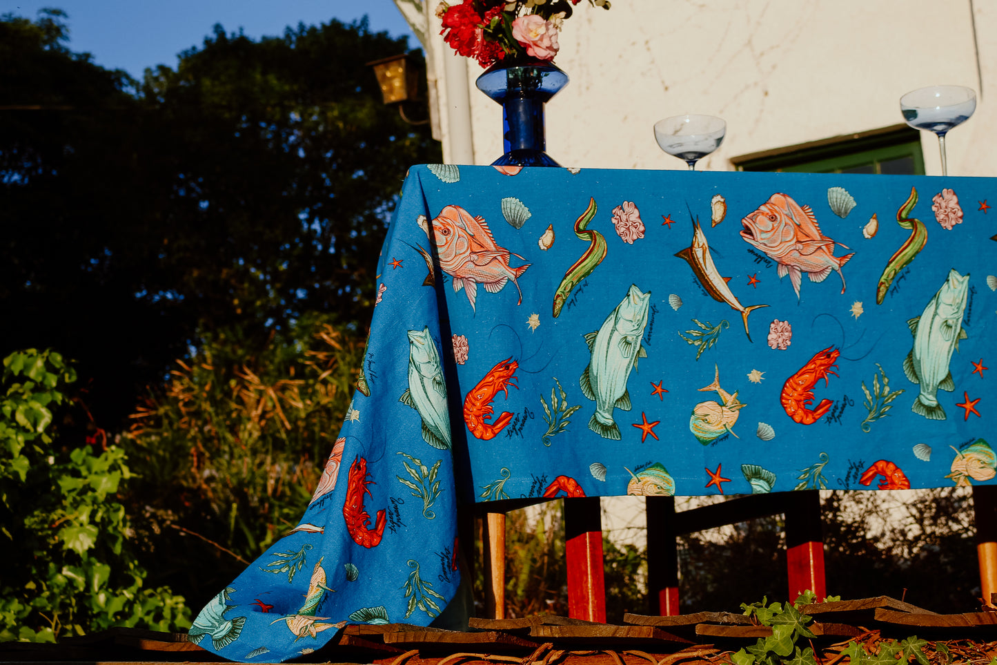 A vibrant funky blue tablecloth titled Fishing for Fishes, adorned with colorful illustrations of fish, lobsters, and seaweed, draped over a table in a sunlit garden setting