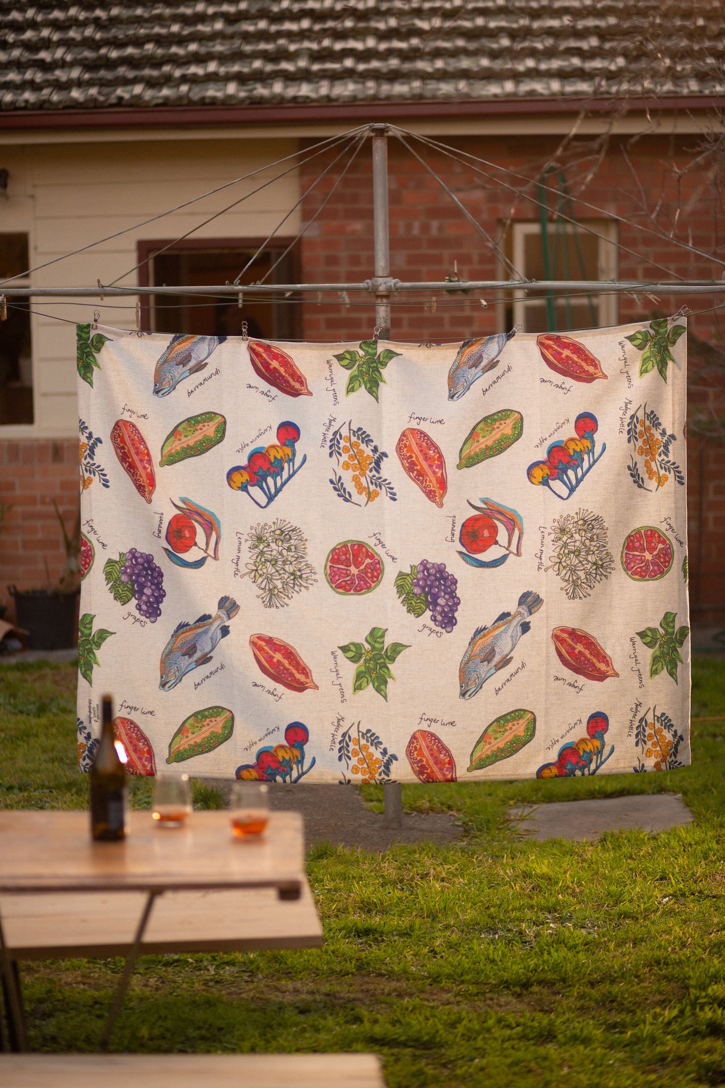 A colorful, illustrated tablecloth with food motifs hanging on a clothesline in a backyard, with a picnic table and drinks in the foreground