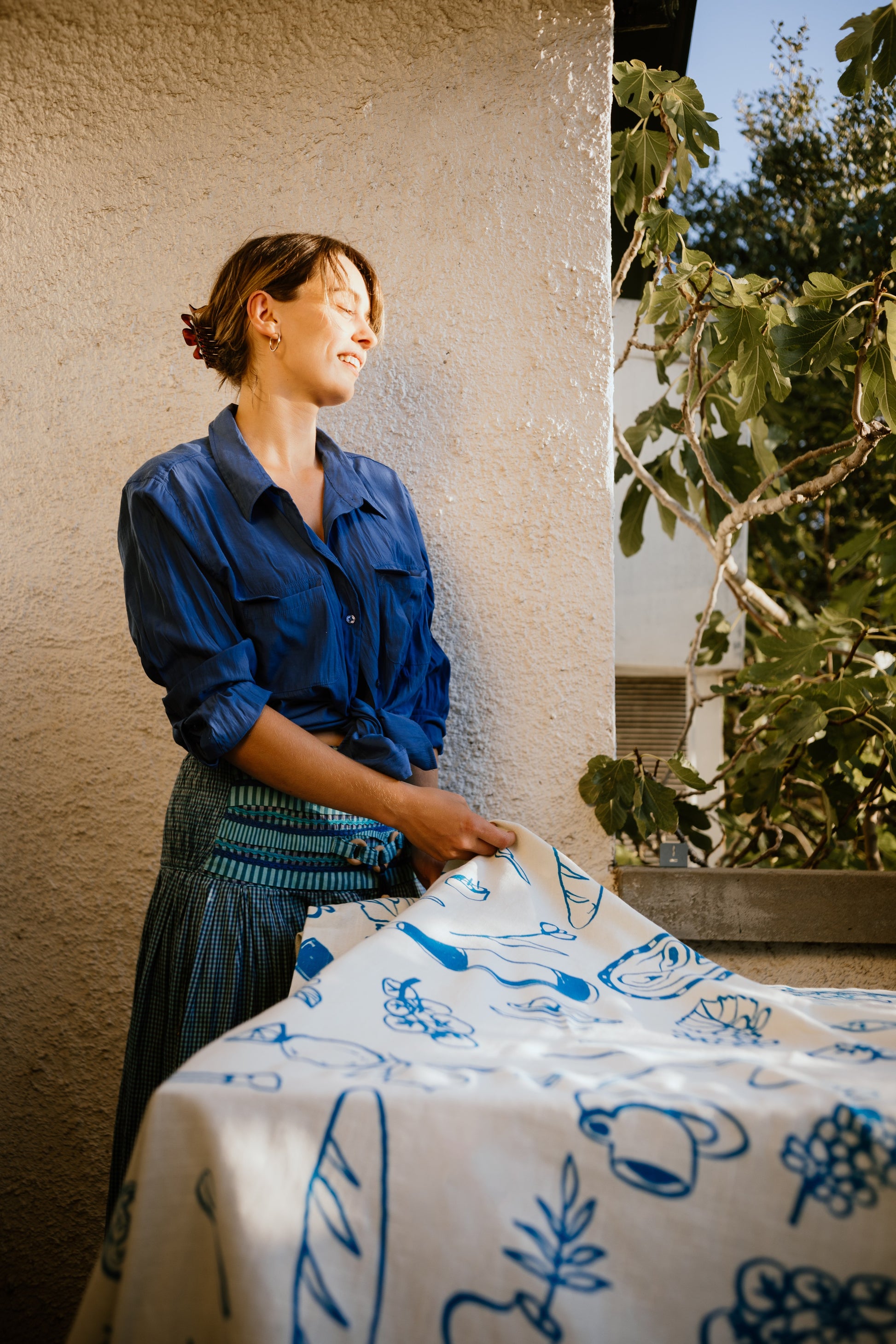 Smiling woman in a blue blouse and striped skirt unfolds a Grecian-themed white and blue tablecloth in a sunlit garden