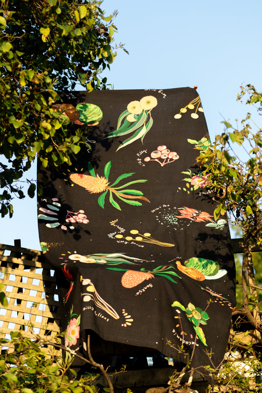 A "Dark Australian Flora" tablecloth hangs against a natural backdrop, its black fabric adorned with vibrant illustrations of Australian plants, showcased outdoors amidst the foliage.