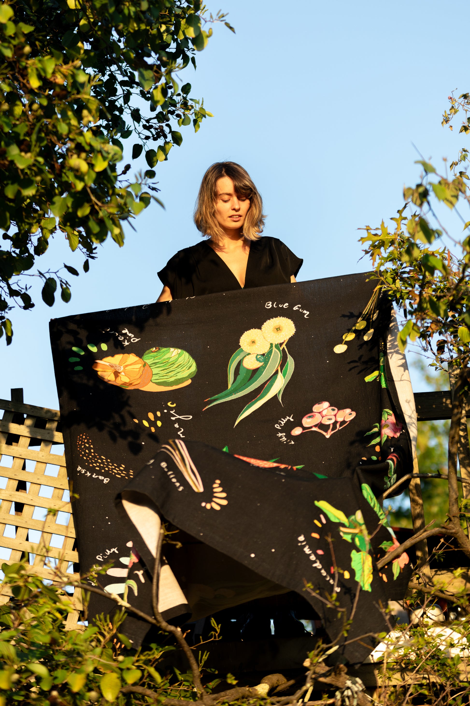 A woman stands holding the "Dark Australian Flora" tablecloth up to the light, its dark backdrop offsetting the bright, colorful depictions of native Australian flora.