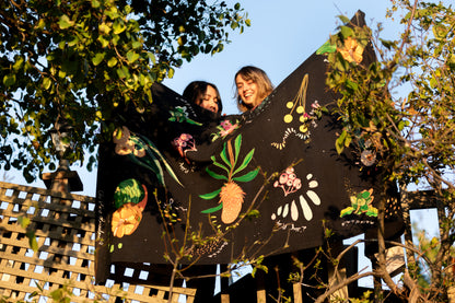 Two women display the "Dark Australian Flora" tablecloth together, revealing intricate designs of Australian native plants. The natural setting emphasizes the connection between the product and the wilderness it celebrates.