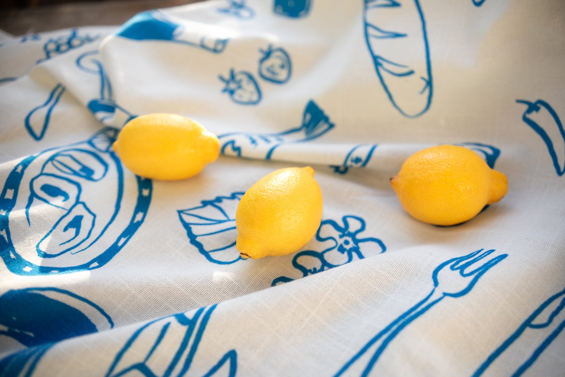 Close-up of a white tablecloth with blue Grecian-inspired prints and fresh lemons resting on top, highlighting the fabric's texture