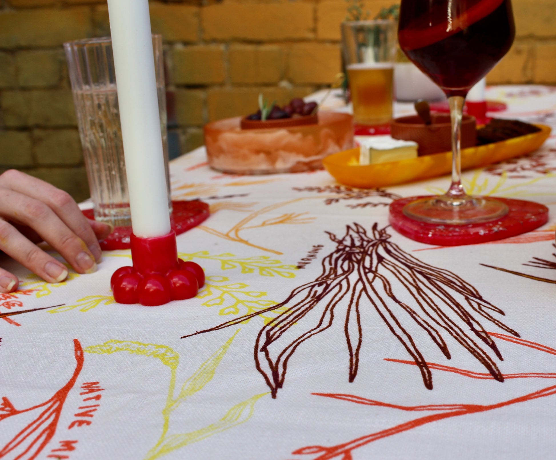 A close-up view of a snack platter on the GRANO tablecloth. The rich orange, yellow, and red patterns of the tablecloth, inspired by the native grains of Australia, provide a vibrant backdrop to bowls of olives, nuts, cheese, and crackers, capturing the warmth and energy of the Australian landscape.