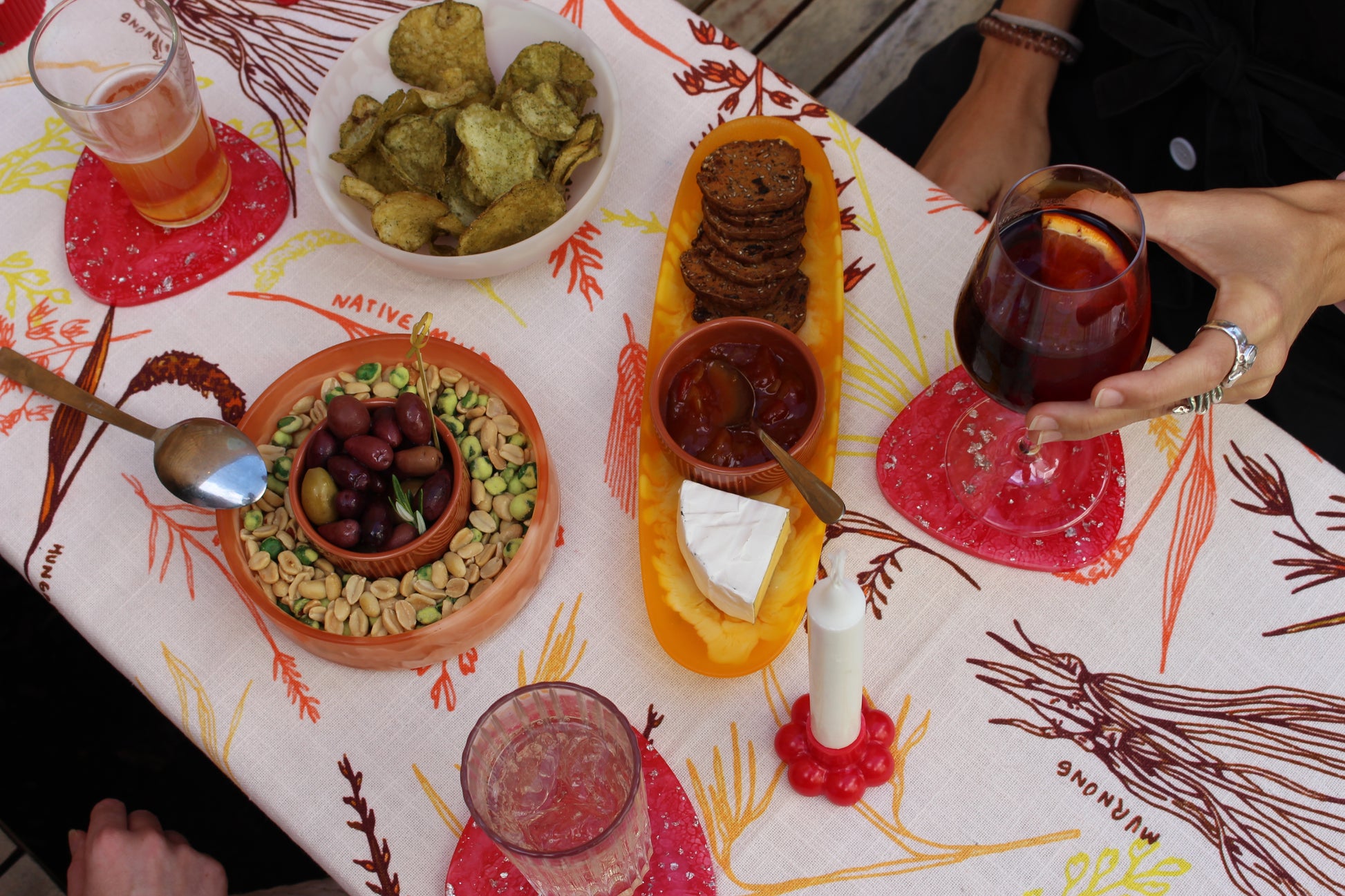 A vibrant table setting with snacks and drinks atop the GRANO tablecloth, showcasing the warmth and vibrancy of the Australian landscape. The rich orange and yellow shades of the tablecloth, inspired by the native grains and the outback, create a lively backdrop for olives, cheese, crackers, and a glass of red wine.