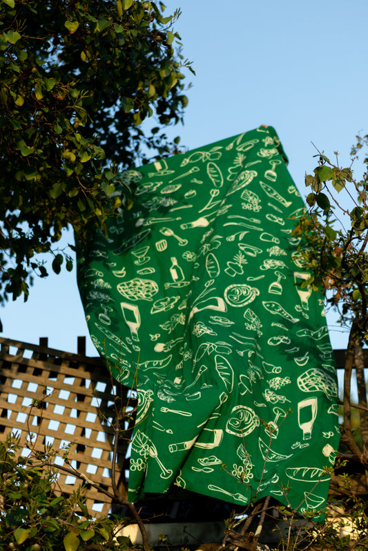 The "Green Feast" tablecloth, inspired by a 2023 summer trip to Greece, draped outdoors, showcasing its bold green hue and simple, elegant design against a backdrop of leafy branches.