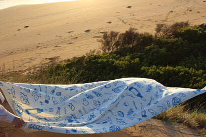 Blue and white patterned tablecloth caught in a mid-air billow against a backdrop of sandy dunes and lush greenery at sunset