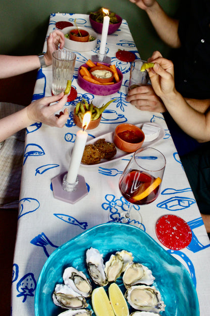 Diners enjoying a meal around a table set with a blue and white Grecian-inspired tablecloth, featuring fresh oysters on a turquoise plate, complemented by various bowls of snacks and glasses of sangria, all illuminated by candlelight.