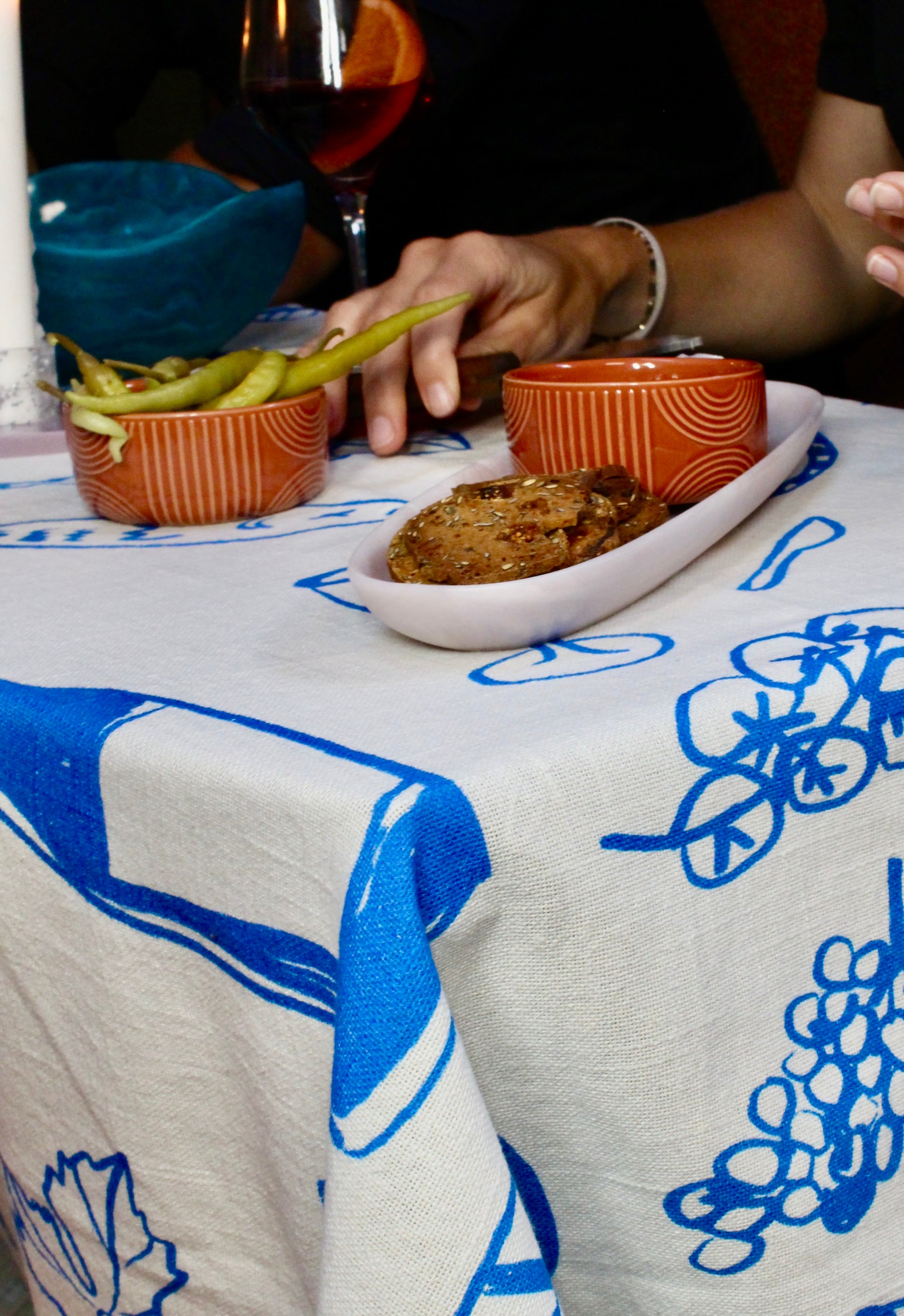 Close-up of a dining table set with a white and blue printed tablecloth, with a hand reaching for a bowl of green olives, next to rustic bread and a glass of wine