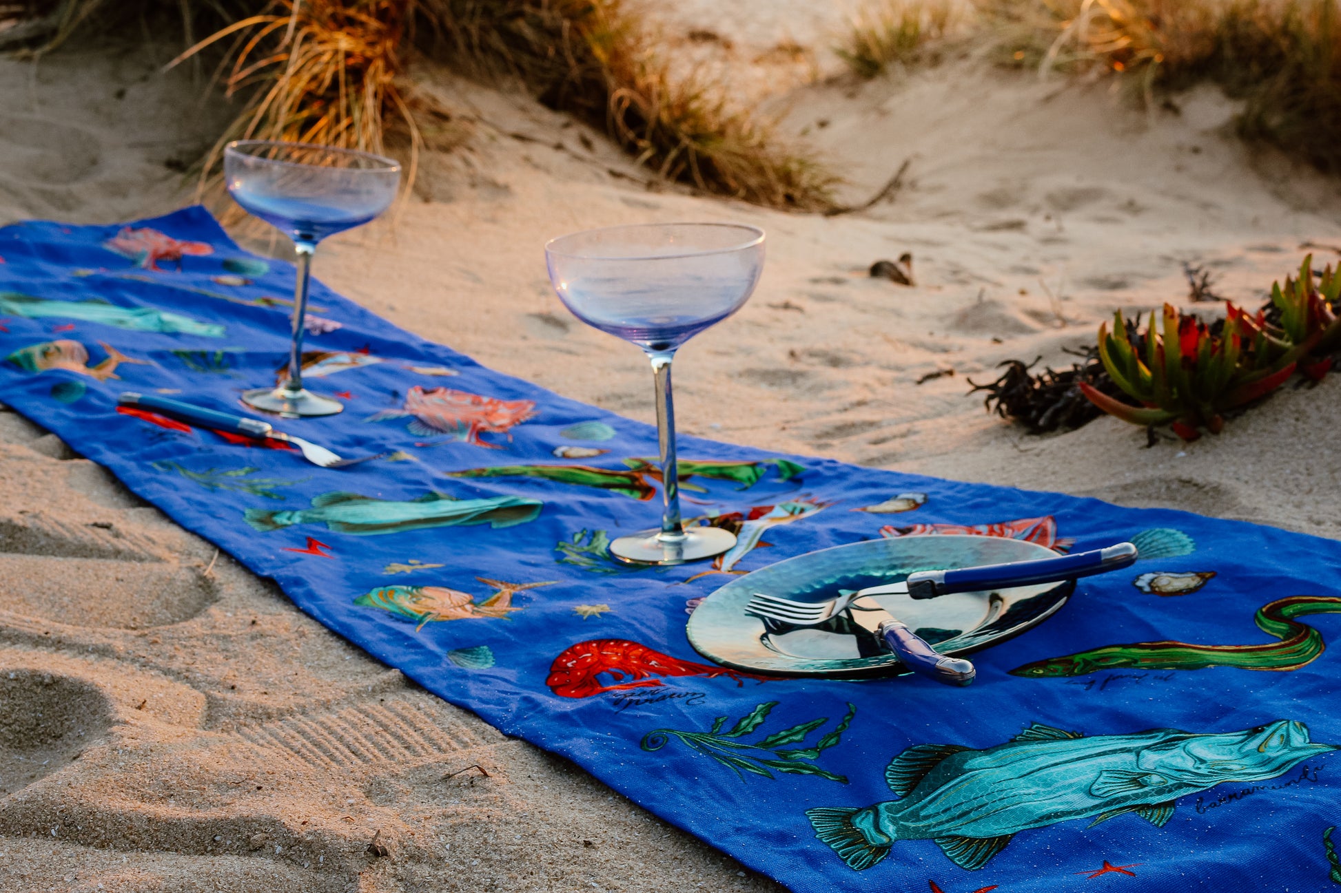 The funky Fishing for Fishes table runner adds a splash of marine magic to the sandy beach, styled with playful blue glassware and vibrant plates, perfect for a whimsical seaside picnic