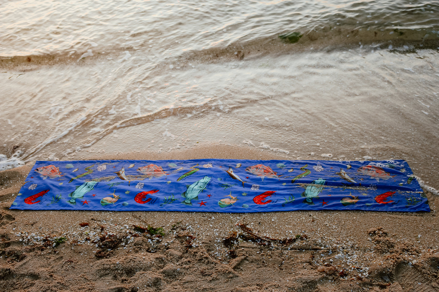 A full-length shot of the Fishing for Fishes table runner stretched along the sand, its bold fish and seaweed patterns popping against the funky coastal backdrop with lively succulents