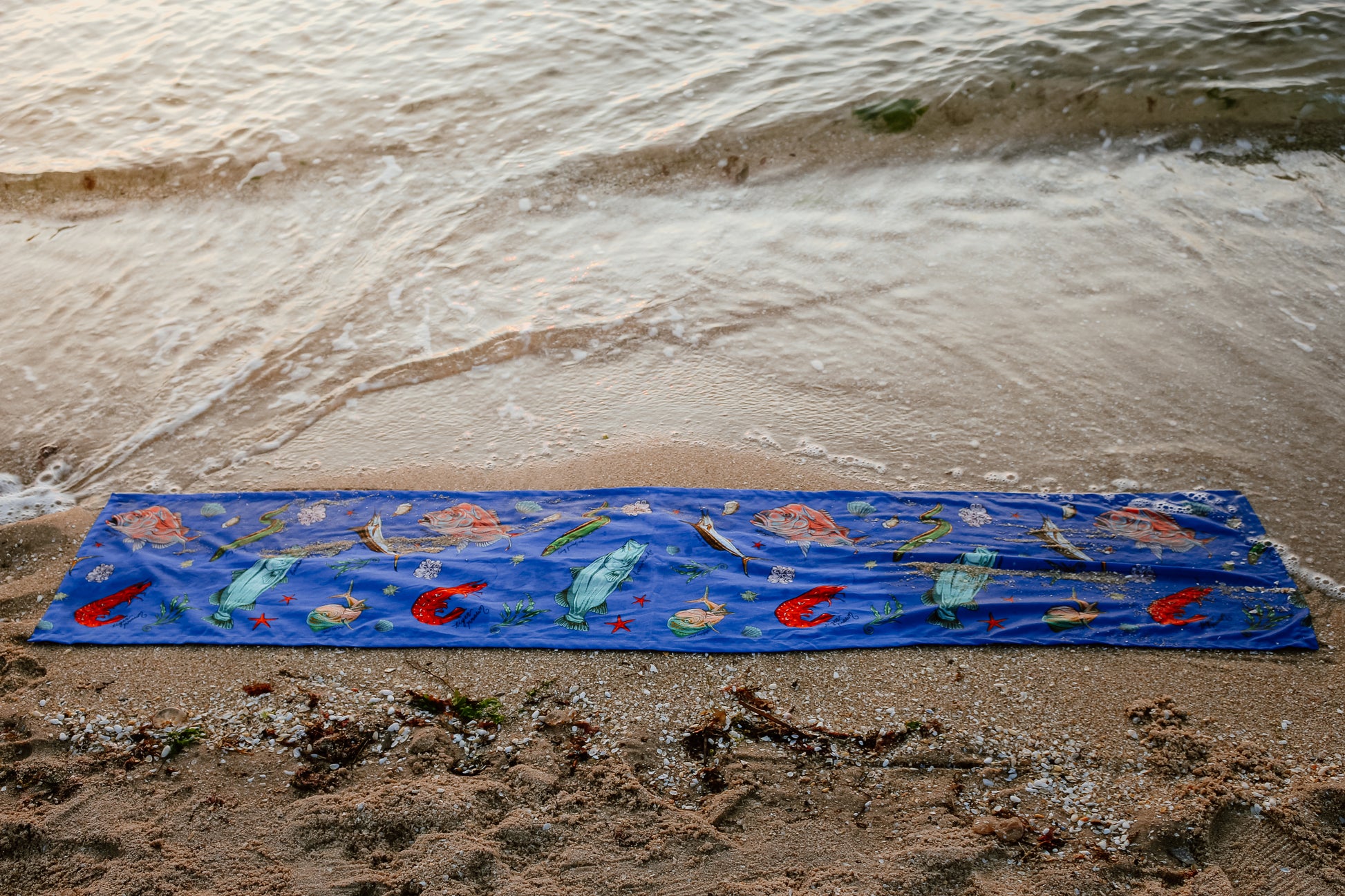 A full-length shot of the Fishing for Fishes table runner stretched along the sand, its bold fish and seaweed patterns popping against the funky coastal backdrop with lively succulents