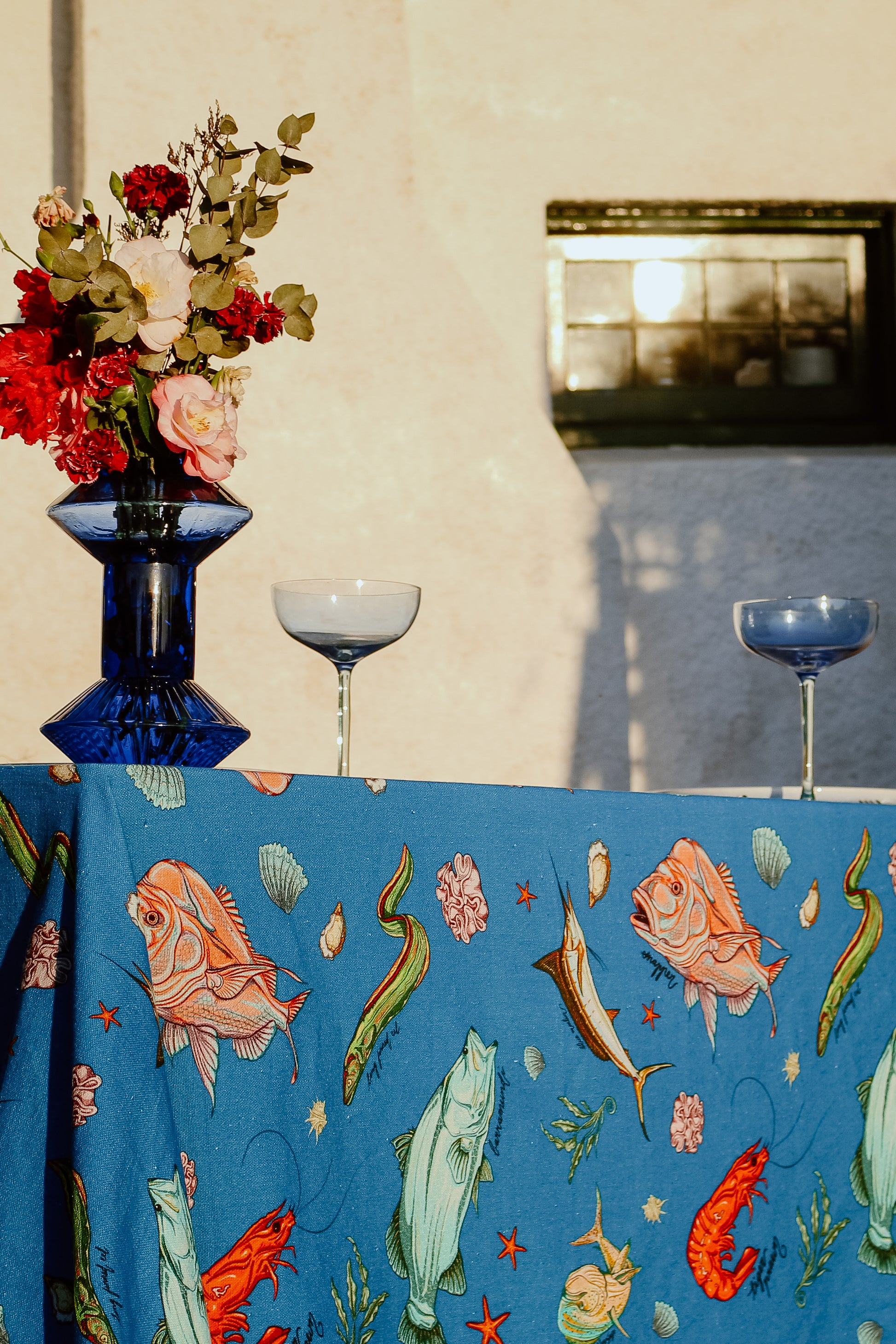 Close-up of the Fishing for Fishes funky tablecloth, showcasing detailed marine designs with a blue vase of fresh flowers and glassware adding a touch of elegance