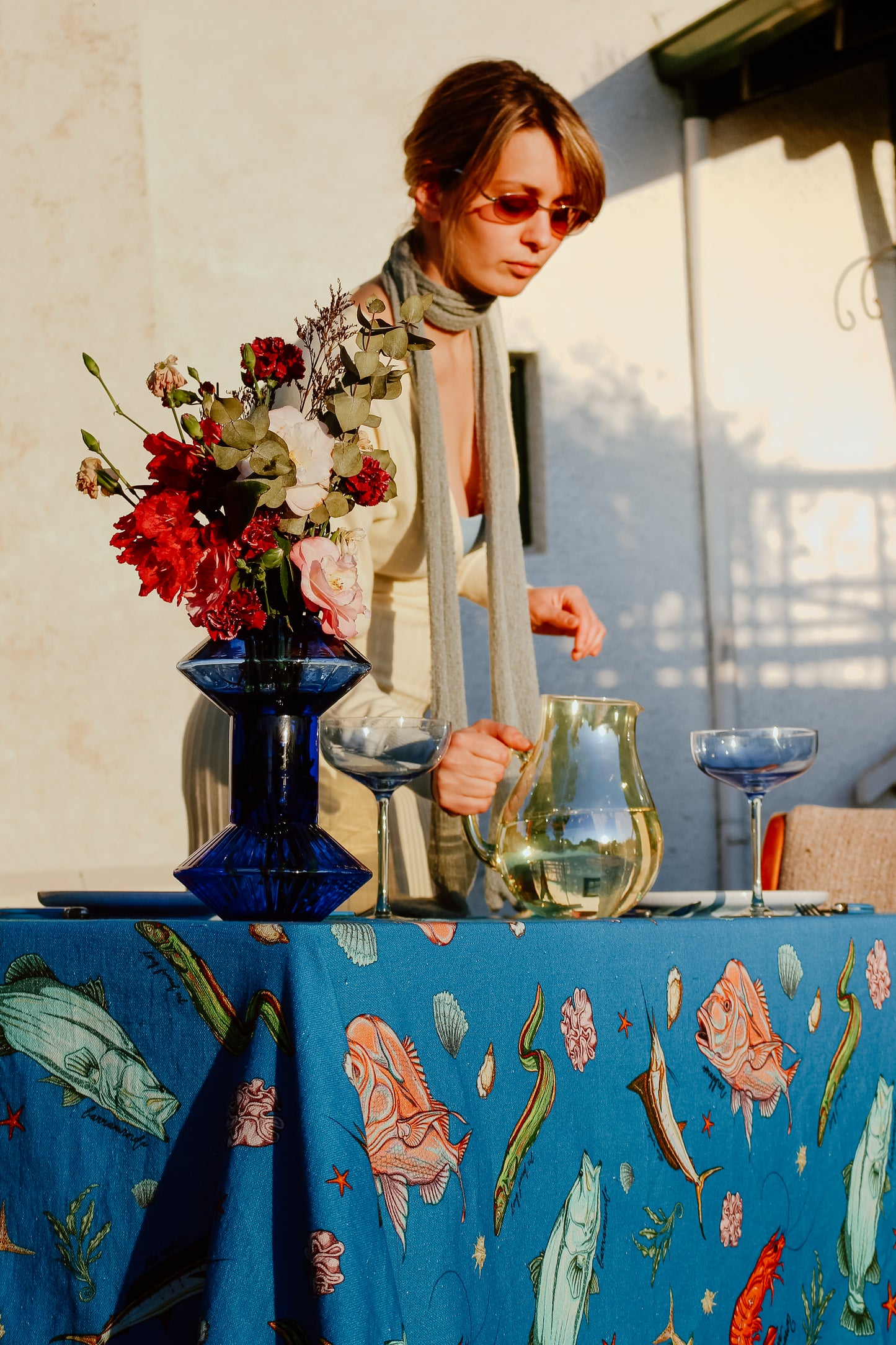 A serene outdoor scene with the Fishing for Fishes funky tablecloth as the centerpiece, styled with a bold blue vase, flowers, and a woman setting the table with a pitcher of water