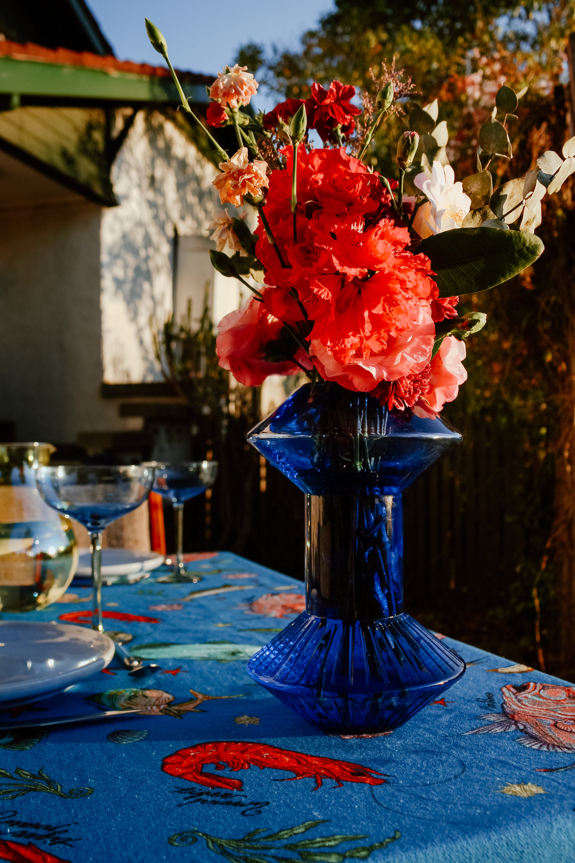 The Fishing for Fishes funky tablecloth under golden sunlight, paired with a striking blue vase filled with bright red and pink flowers, creating a cheerful and inviting tablescape