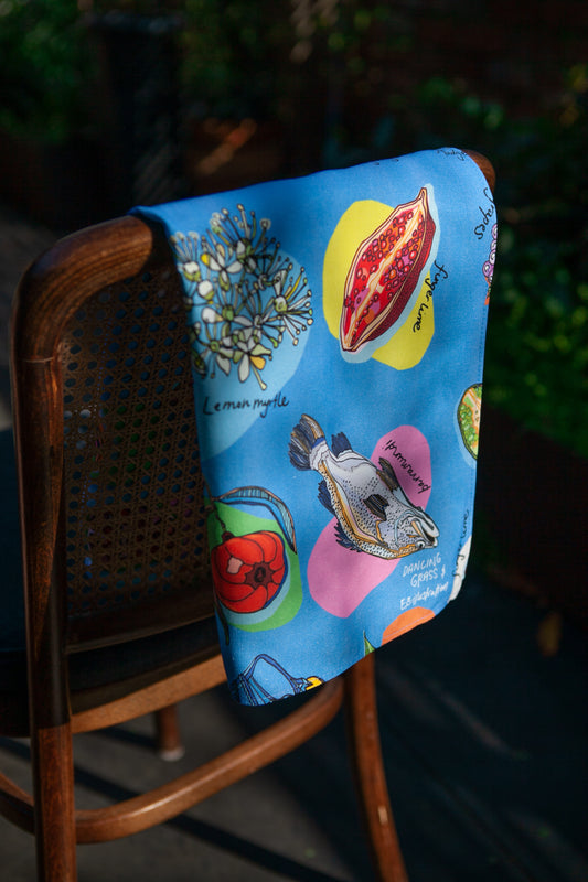 Artistically designed white tea towel with colorful fruit and herb prints, draped over the back of a wooden chair.