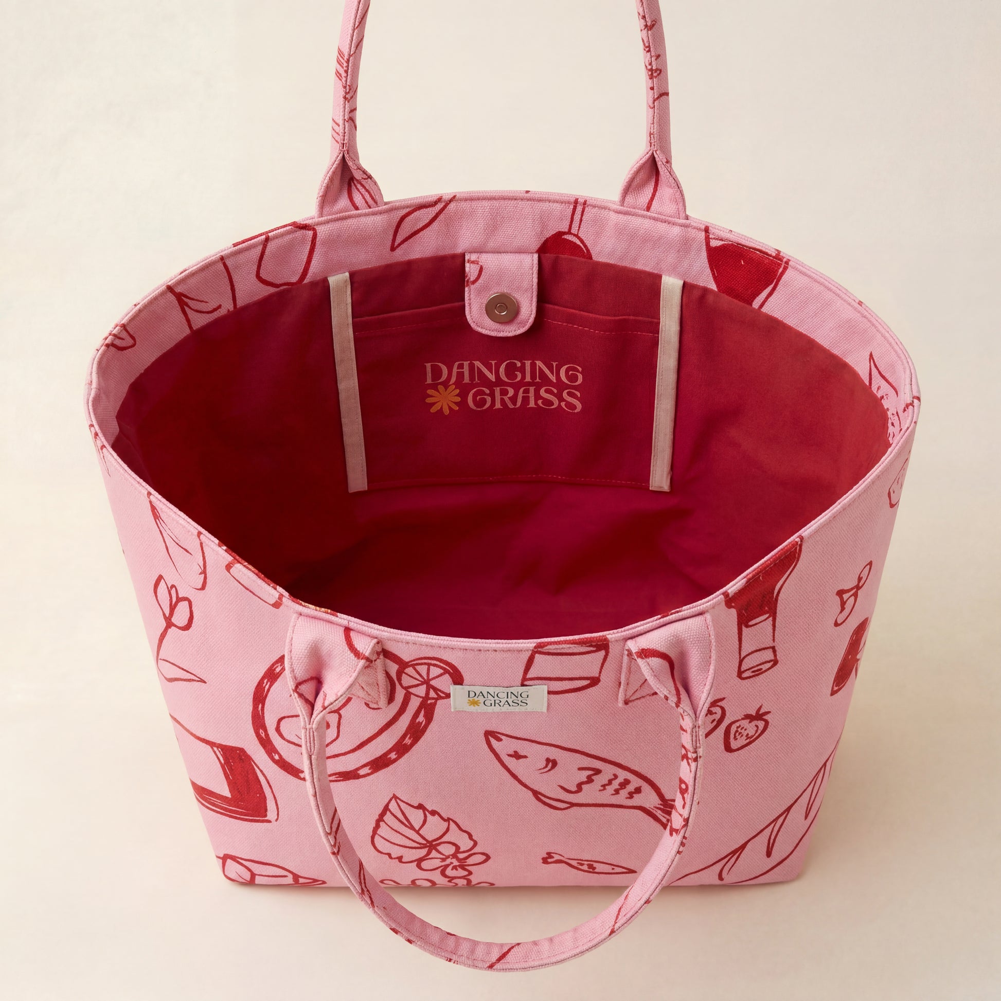 Pink tote bag with red interior and Dancing Grass branding on a beige background