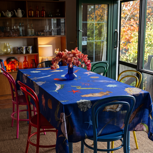 Dining room with australian fish tablecloth, colorful chairs, and a window view of trees.