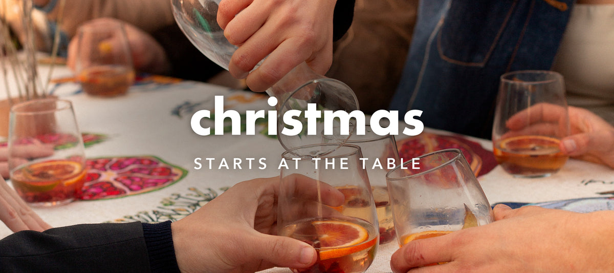 Friends pouring blood orange drinks over the Dancing Grass Home Grown tablecloth, with the words Christmas starts at the table