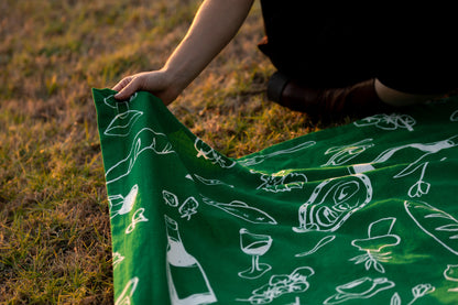 A close-up shot of a hand gently holding the edge of the Green Feast tablecloth. The vibrant green fabric features minimalist white illustrations inspired by a 2023 summer trip to Greece, reflecting the elegance and simplicity of its design.