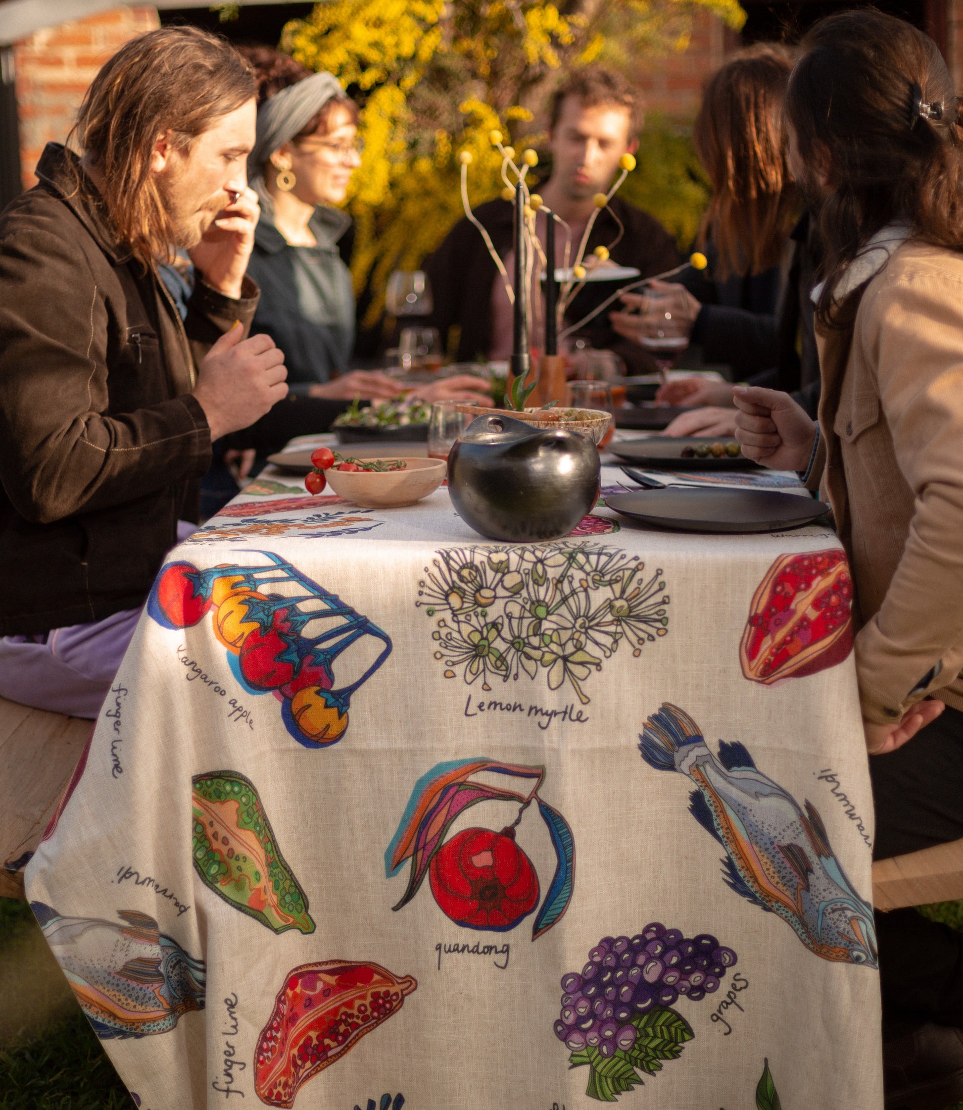 A lively scene around a dining table where guests enjoy food and drinks while admiring the artistic details of the 'Home Grown' tablecloth, featuring native bush food illustrations like lemon myrtle, finger lime, and quandong.