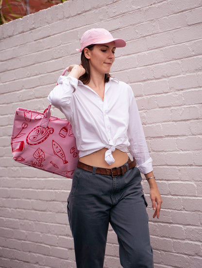 Pink Feast tote bag worn over the shoulder by a woman in a white tied shirt and pink cap
