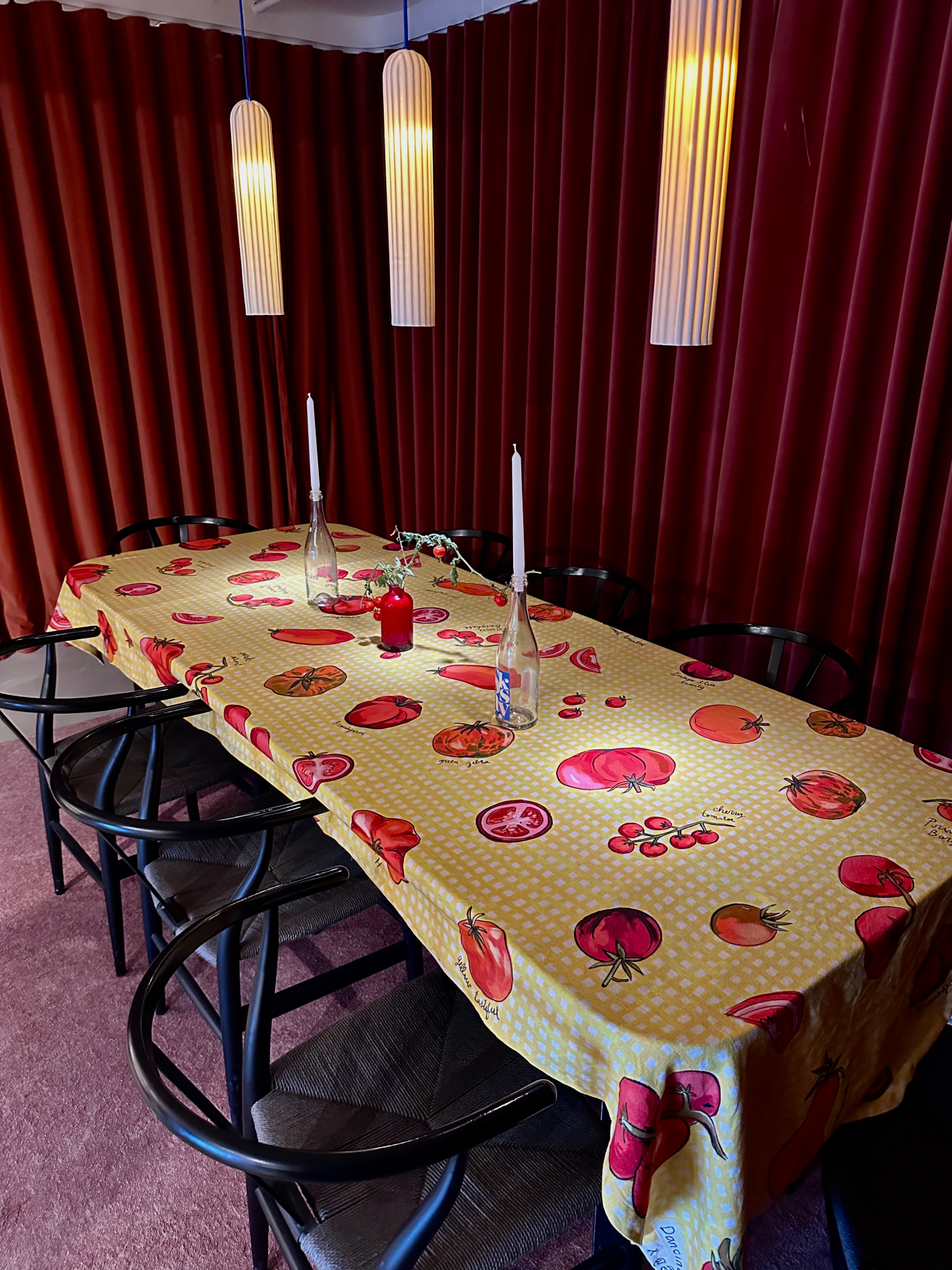 Long dining table set with the Red Juicy Mess tomatoe tablecloth. Red velvet curtains in the background, soft candlelight above, and tomatoes illustrated across the cloth.