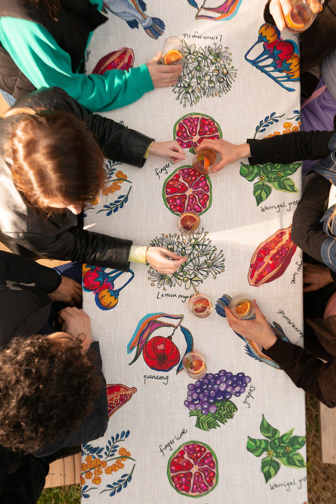 Hands reach across a dining table set with drinks and food, as guests point out and admire the unique native food illustrations, such as grapes and lemon myrtle, on the beautifully designed tablecloth.