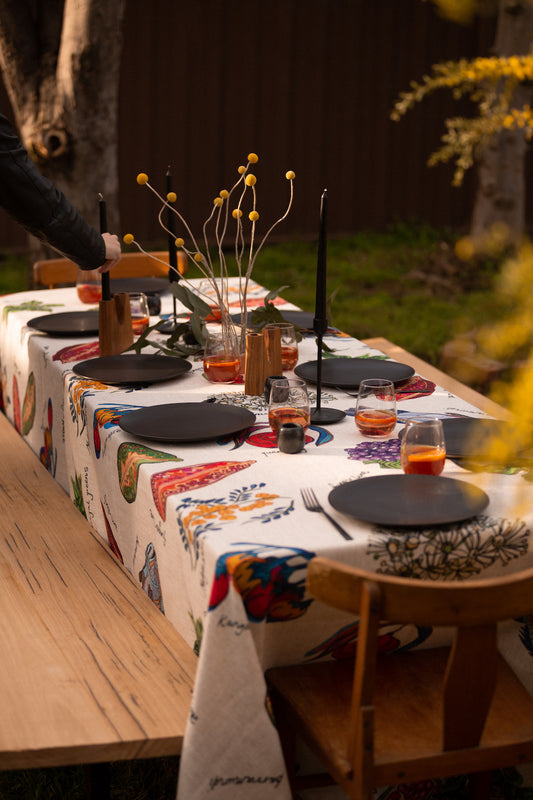 A beautifully set outdoor dining table features a vibrant Australian-designed tablecloth adorned with illustrations of native bush foods, while someone serves a stunning dessert topped with native berries.